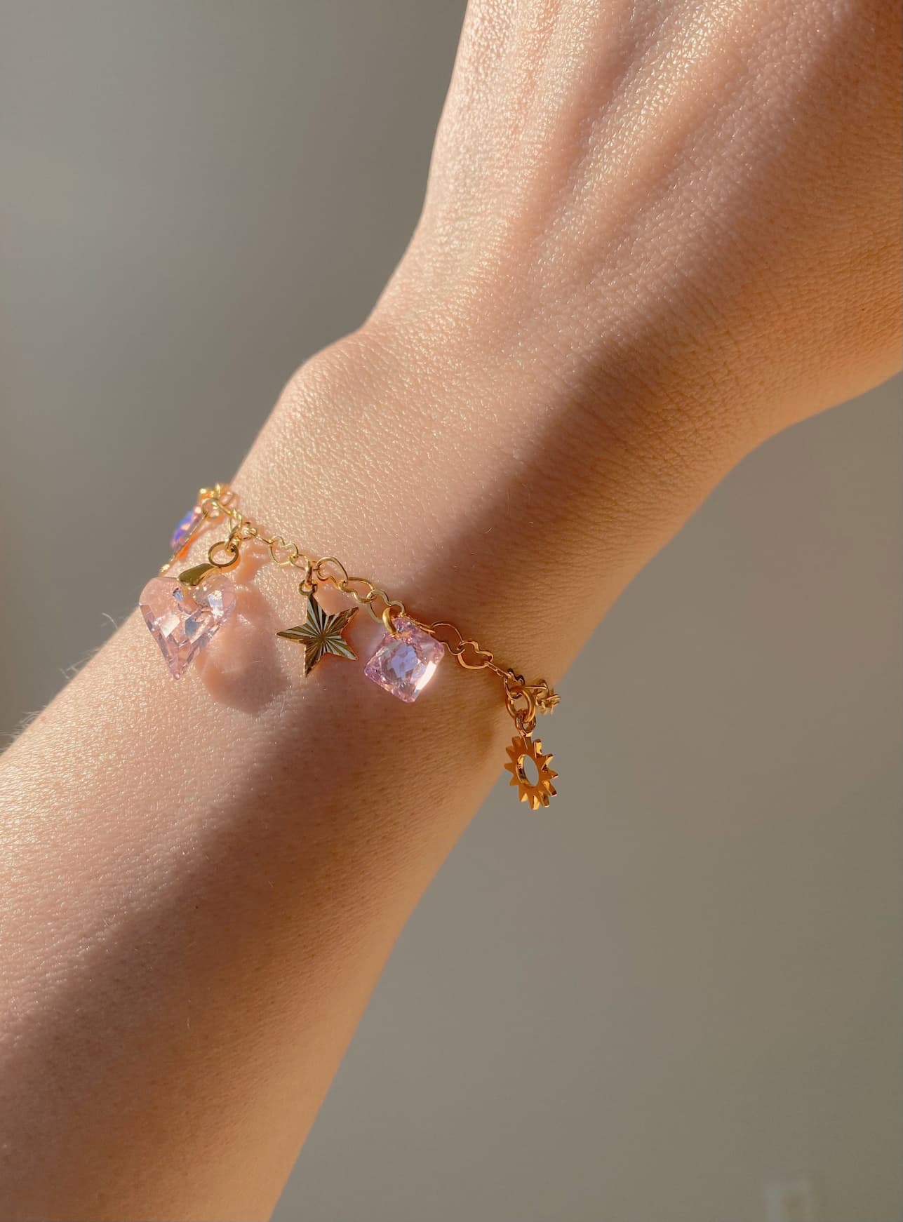 A hand adorned with the exquisite Henriette Bracelet by Noémiah, featuring a delicate gold heart chain and small charms like a star and a sun. The bracelet is beautifully enhanced with pink, crystal-like Swarovski crystals. The softly lit background elegantly highlights the jewelry's intricate details.