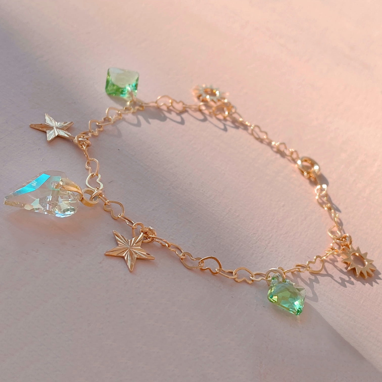 The Henriette Bracelet by Noémiah, a delicate gold-plated piece featuring star and sun charms accented with green and faceted Swarovski crystals, gracefully rests on a soft pink surface illuminated by natural light.