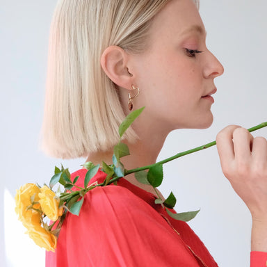 A woman with short blonde hair holds a yellow rose. In profile view, she wears a red top and the Huguette Earrings by Noémiah, which are heart-shaped and adorned with Swarovski crystals. The lightweight earrings gleam against the soft, neutral backdrop.