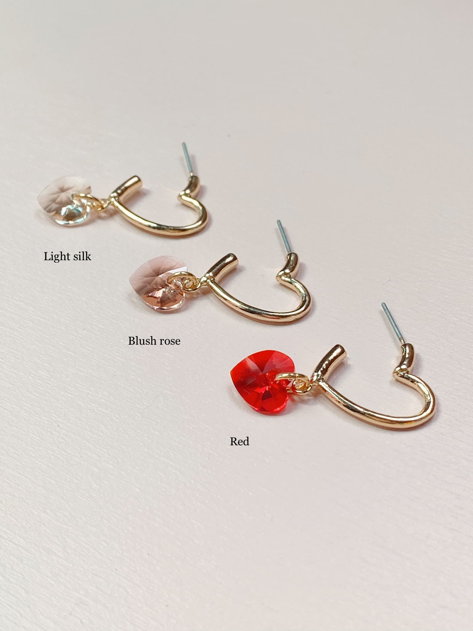 The Huguette Earrings by Noémiah are gracefully arranged on a white surface, featuring three heart-shaped earrings with gold hoops. Each lightweight piece showcases a heart charm in varying colors: light silk, blush rose, and red. Embellished with dazzling Swarovski crystals and resistant to tarnish, the earrings are labeled by color above each one for easy identification.