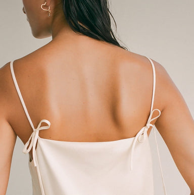 A person with long dark hair is seen from the back, wearing a beige spaghetti strap dress featuring bow details. They are also accessorized with the lightweight and tarnish-resistant Huguette Earrings by Noémiah, which are heart-shaped. The background is plain and neutral.