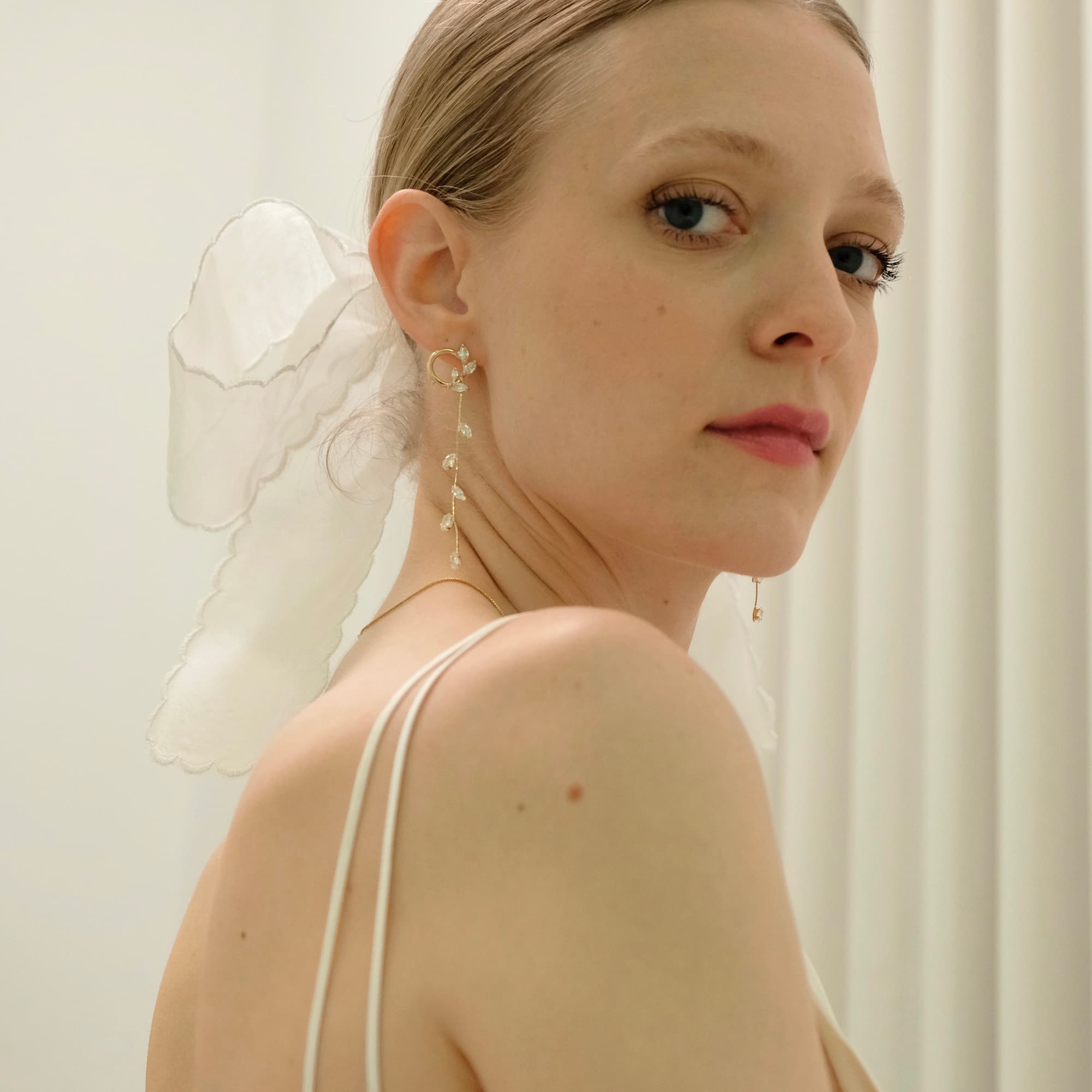 A woman with light hair tied back with a sheer white ribbon glances over her shoulder. She wears Noémiah's Inès Earrings, featuring gold-plated hoops, along with a thin-strapped top set against a softly lit background adorned with white drapery.