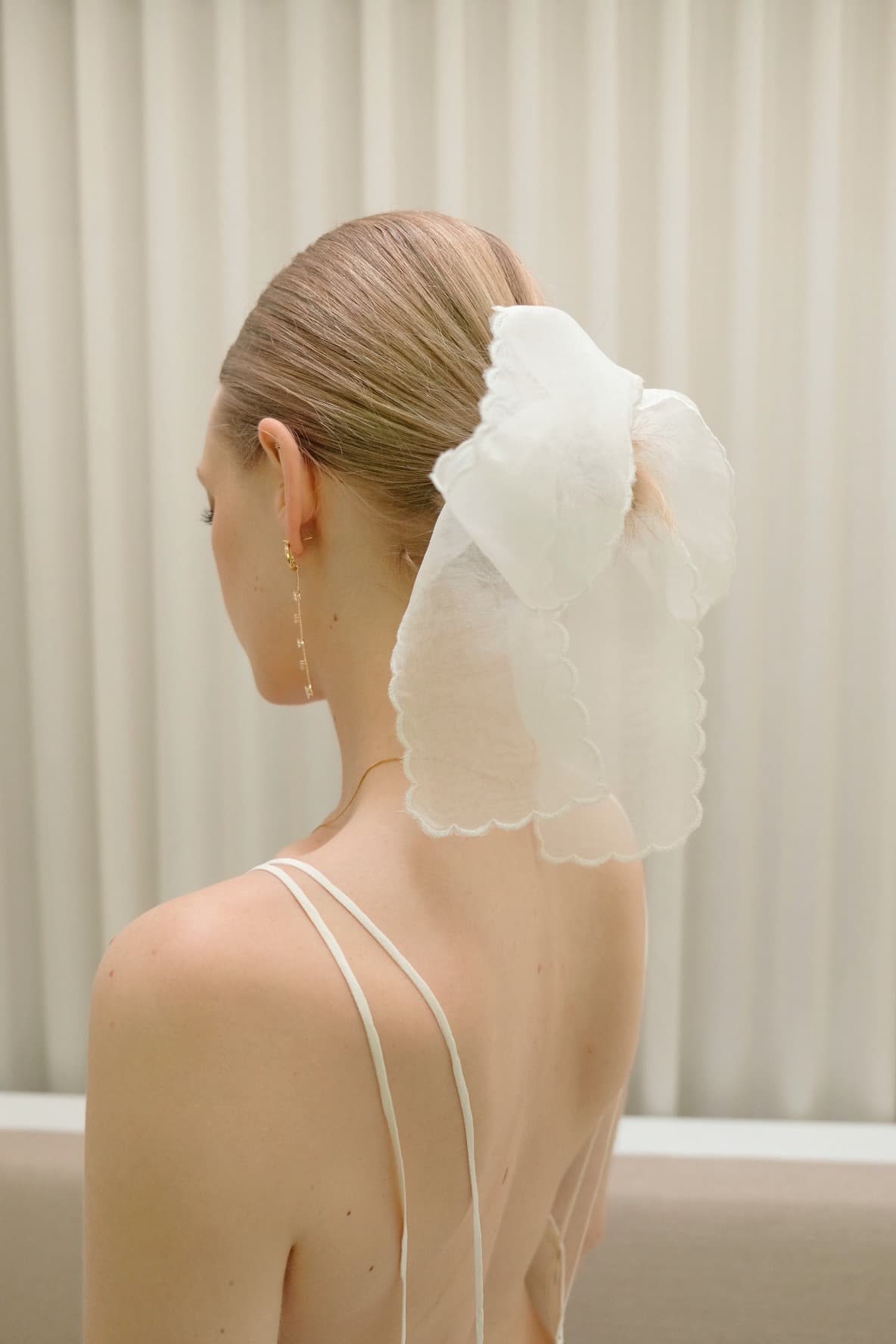 A woman with light brown hair styled in a bun, adorned with a large white bow. She is wearing a spaghetti strap dress and the elegant Noémiah Inès Earrings. The background features white curtains.