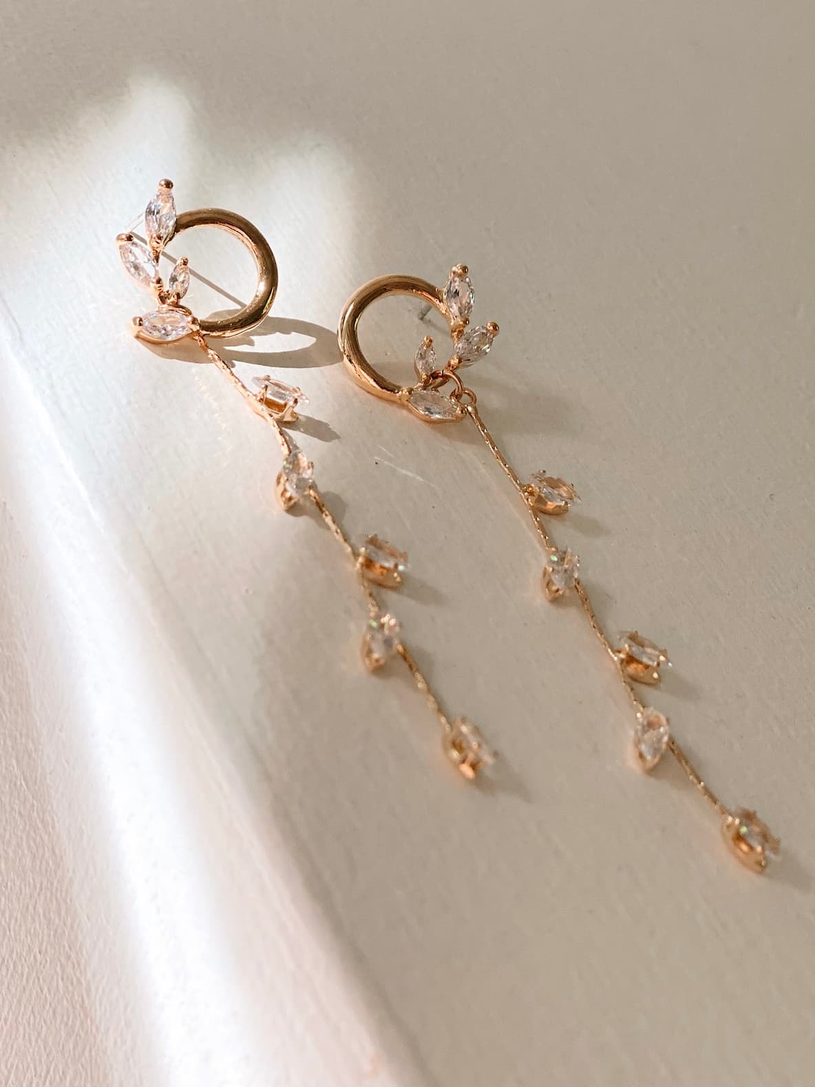 The Inès Earrings by Noémiah, featuring elegant gold-plated hoops with delicate dangling chains and clear crystal accents, are showcased on a light surface bathed in soft, natural light.