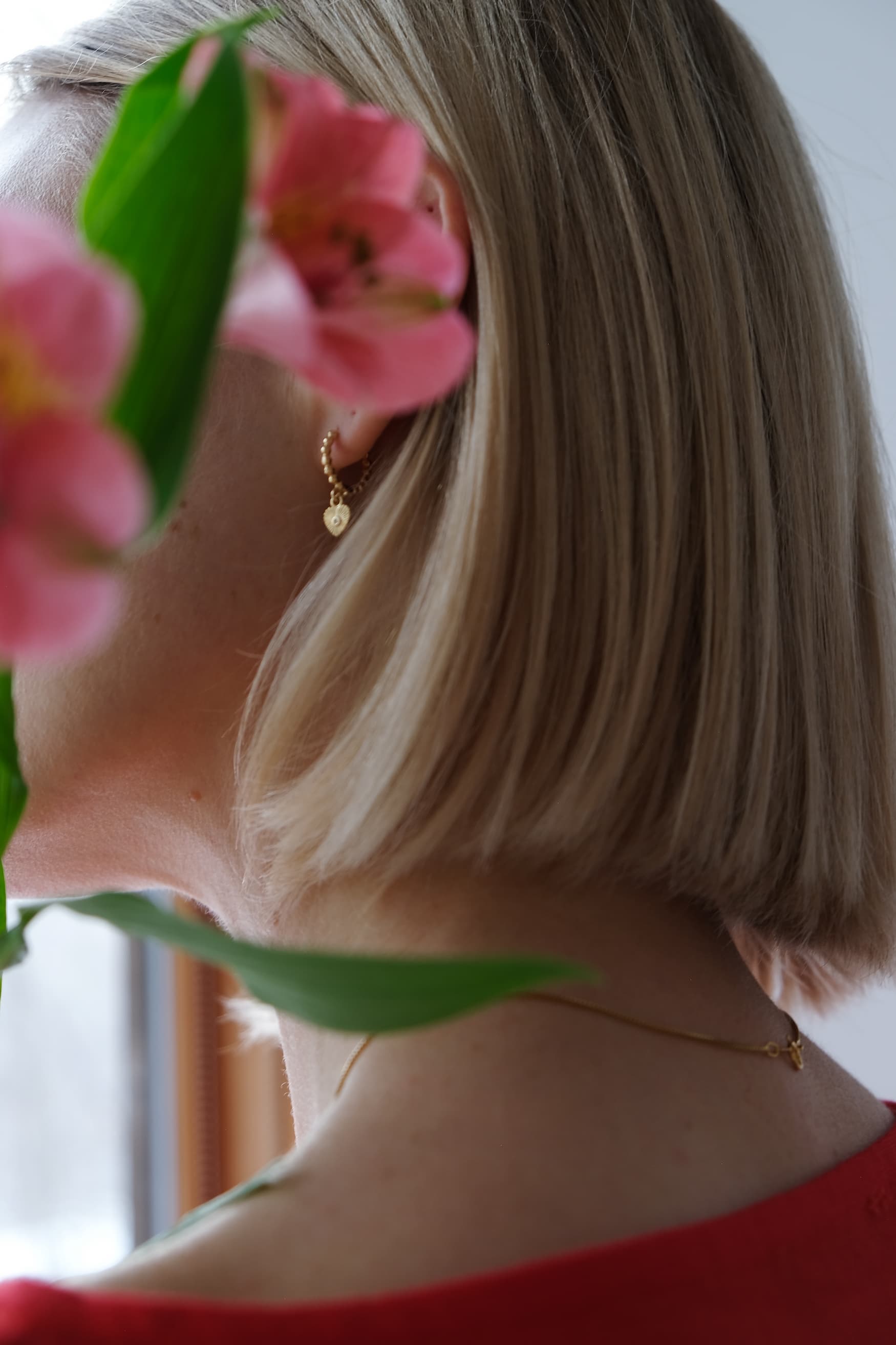 A person with short blonde hair is partially obscured by pink flowers, wearing a gold chain necklace and the shimmering Ingrid Earrings from Noémiah. The background appears to be indoors near a window.