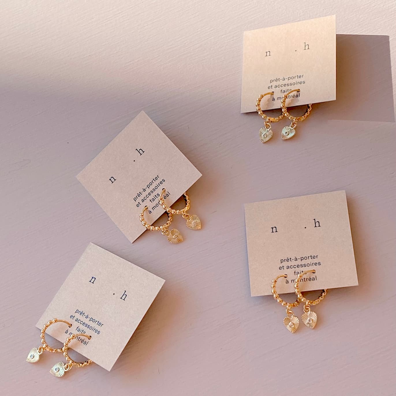 Four pairs of the Ingrid Earrings by Noémiah, crafted in gold plate with small white charms, are showcased on individual beige cards. Each card highlights branding and text about prêt-à-porter and accessories from Montreal. Under soft, natural light, these delicate pieces shimmer gracefully with cubic zirconia accents.