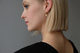 A side profile of a person with short blonde hair is adorned with the Ingrid Earrings by Noémiah and wearing a black shirt, set against a plain gray background.