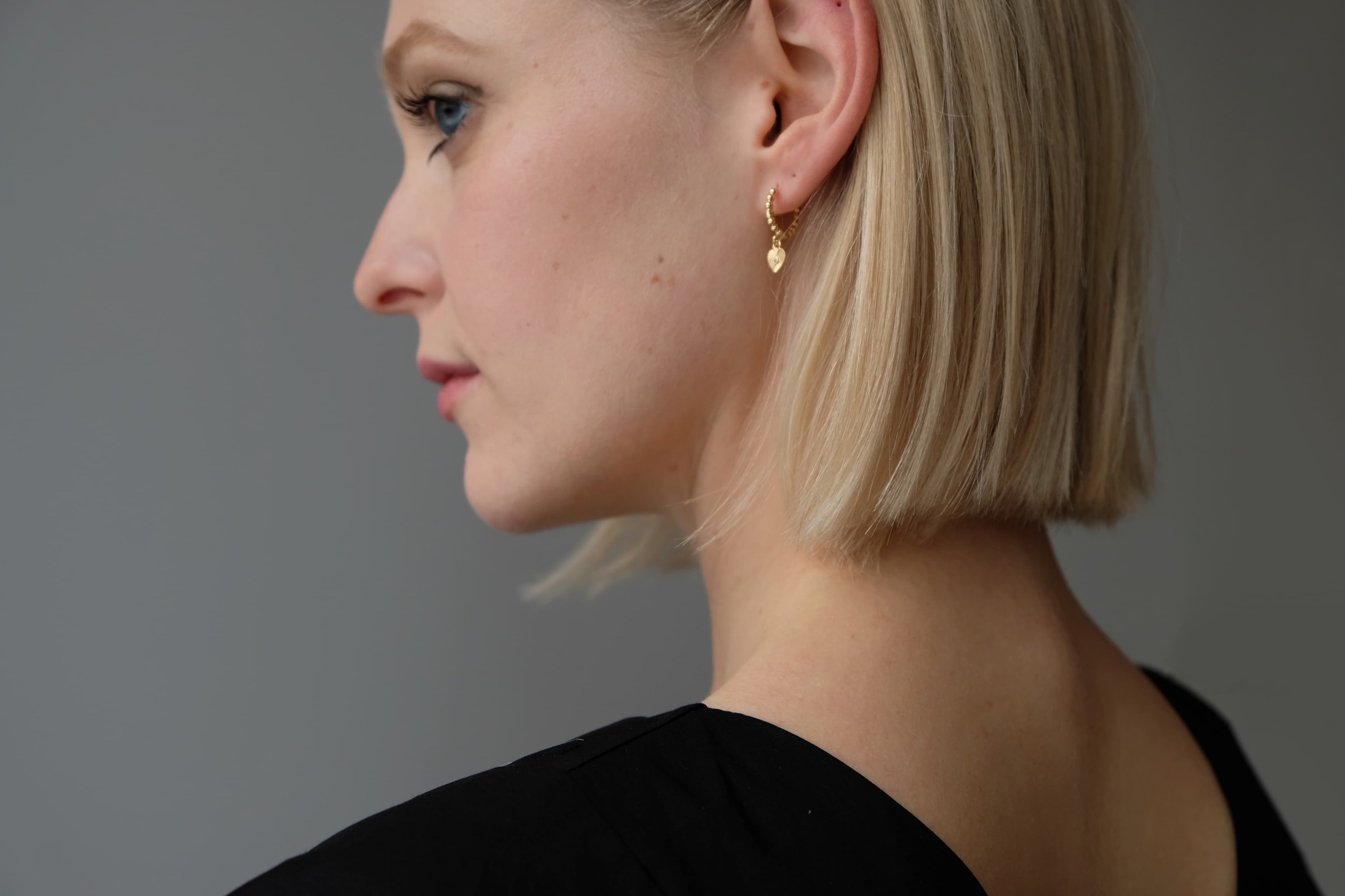 A side profile of a person with short blonde hair is adorned with the Ingrid Earrings by Noémiah and wearing a black shirt, set against a plain gray background.