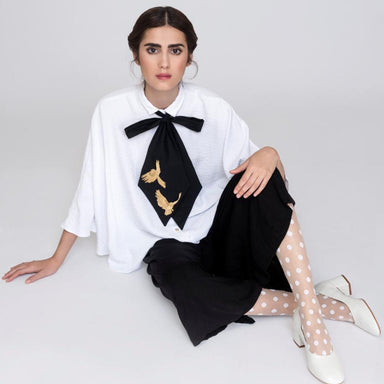 A woman sits gracefully on the floor in an elegant white blouse featuring Noémiah's Iris Necktie, embellished with a large black bow adorned with gold bird designs. She pairs it beautifully with a black skirt, white polka dot stockings, and white heels against a light background.