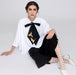 A woman sits gracefully on the floor in an elegant white blouse featuring Noémiah's Iris Necktie, embellished with a large black bow adorned with gold bird designs. She pairs it beautifully with a black skirt, white polka dot stockings, and white heels against a light background.