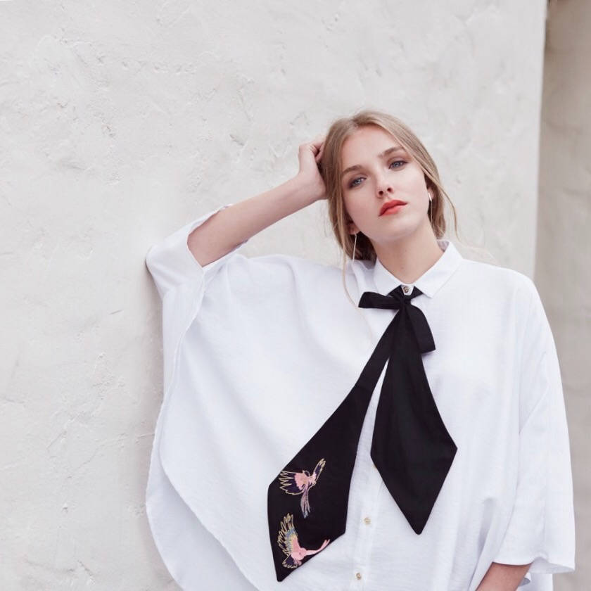 A woman wearing a wide-sleeved silk cotton shirt and the Iris Necktie from Noémiah, featuring embroidered birds, stands against a light-colored wall. She rests one arm on her head and complements her look with subtle makeup and red lipstick.