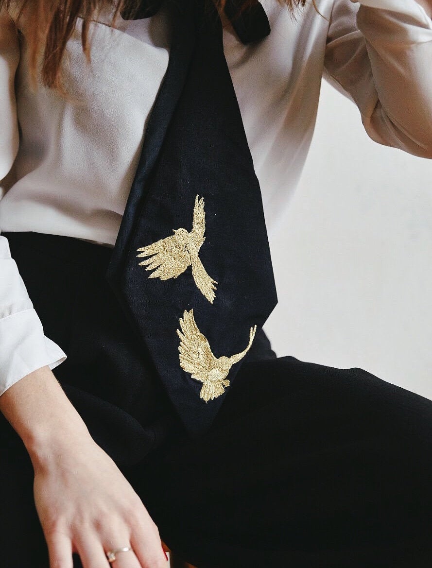 A person wearing a white shirt and the Iris Necktie by Noémiah, a handmade black tie featuring two embroidered gold birds. This women's necktie is prominently displayed against black pants, and the person's left hand is resting on their lap.