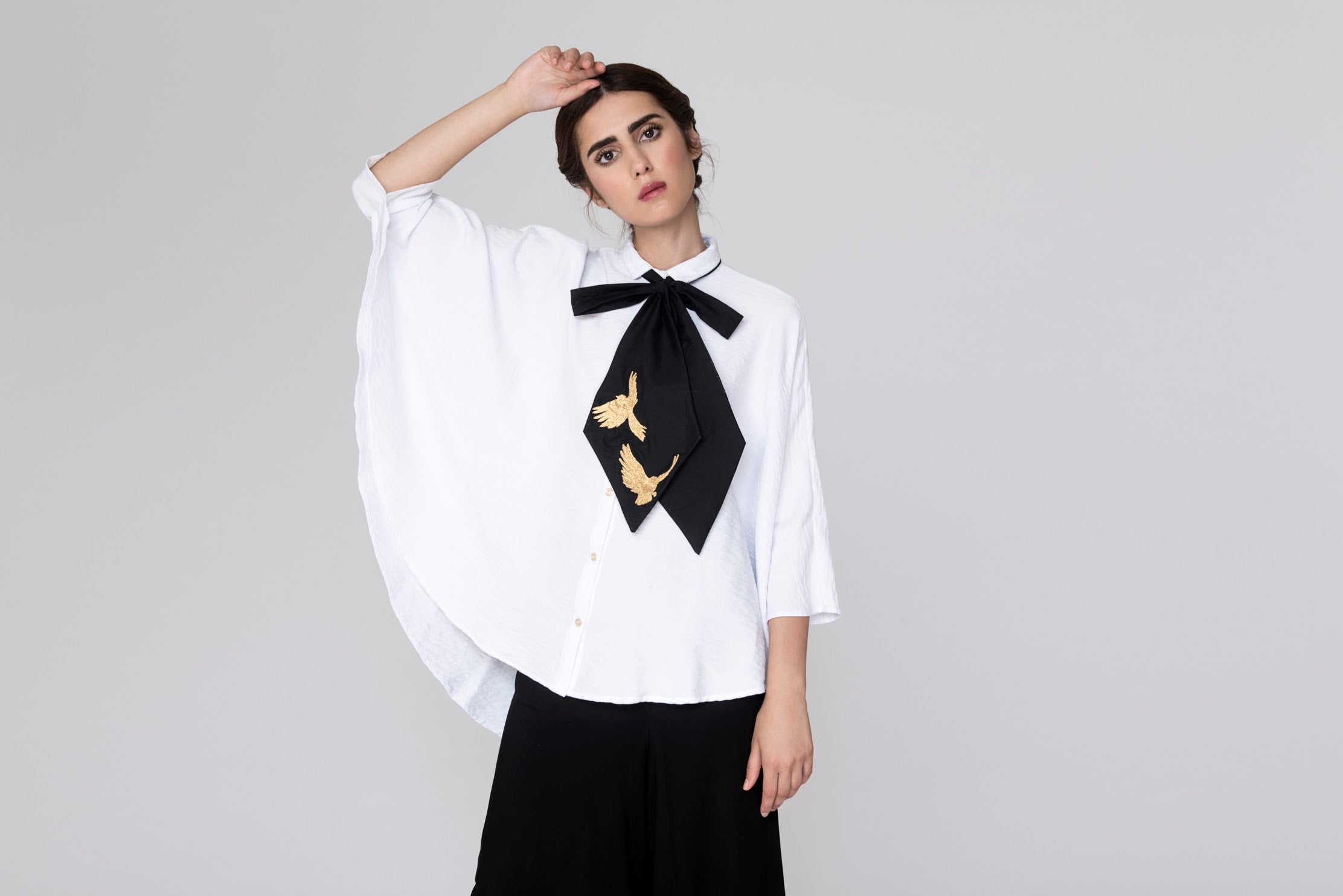 A person stands against a plain background, wearing the chic Iris Necktie blouse from Noémiah. This handmade piece, crafted from silk cotton, features a substantial black bow adorned with elegant gold bird designs and a unique cape-like sleeve on one side. The person gazes directly at the camera with one hand resting gently on their head.