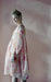 Set against a soft pink background, a person stands elegantly in a flowy Céline Floral Dress by Noémiah, adorned with a floral pattern that calls to mind an Emily Dickinson herbarium. With their light hair styled in a bun and clutching a small lace accessory, the lighting enhances the dreamy ambiance.