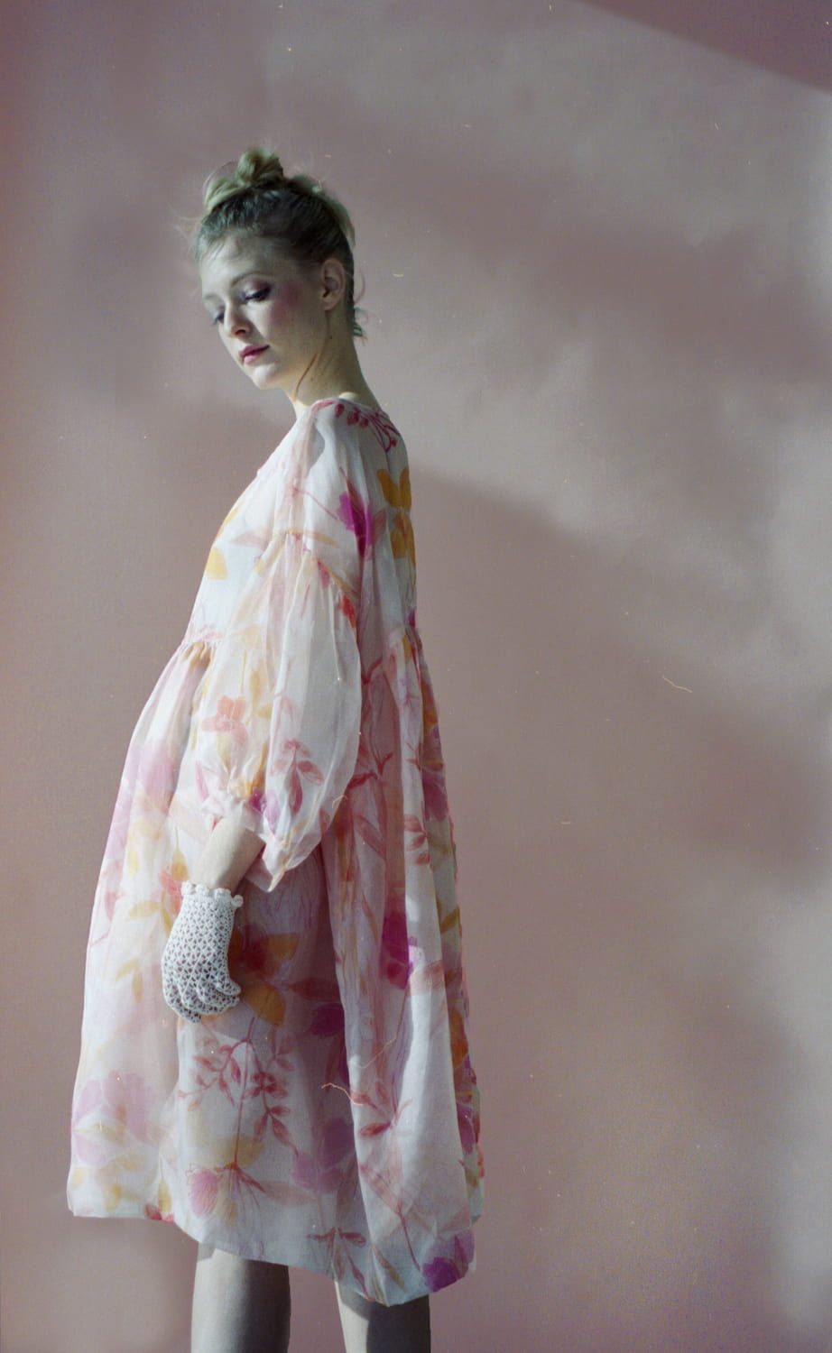 Set against a soft pink background, a person stands elegantly in a flowy Céline Floral Dress by Noémiah, adorned with a floral pattern that calls to mind an Emily Dickinson herbarium. With their light hair styled in a bun and clutching a small lace accessory, the lighting enhances the dreamy ambiance.