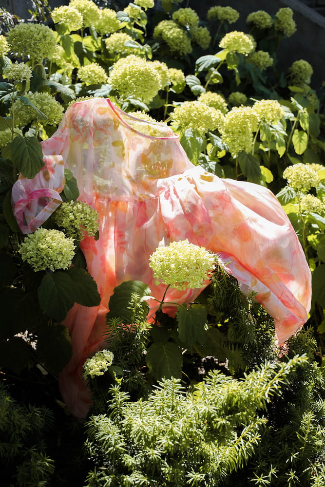 A Noémiah Céline Floral Dress, adorned with pastel hues of pink and orange, elegantly drapes over green hydrangea bushes in full bloom. The silk organza offers a vibrant contrast against the rich garden foliage, evoking the charm of an Emily Dickinson herbarium.