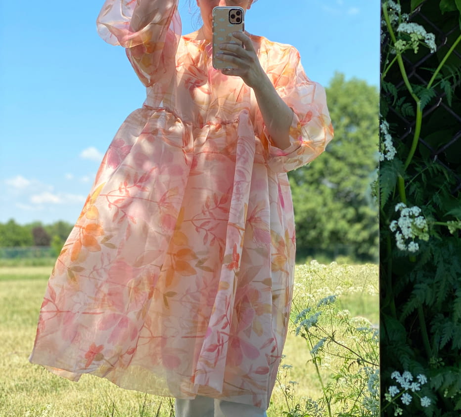 A person holding a phone captures a photo of themselves outside in the grassy field under a clear blue sky. They are wearing a flowing Céline Floral Dress by Noémiah, featuring pink and orange floral patterns reminiscent of an Emily Dickinson herbarium.