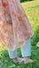 A person stands barefoot on green grass, clad in a pink and orange Céline Floral Dress by Noémiah that resembles silk organza. The sunlit field behind them blooms with plants and flowers, evoking the essence of an Emily Dickinson herbarium.