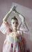 A person is wearing a Céline Floral Dress by Noémiah with sheer puff sleeves crafted from silk organza and lace gloves. Their arms are raised above their head, reminiscent of an Emily Dickinson herbarium, against a soft pink background.