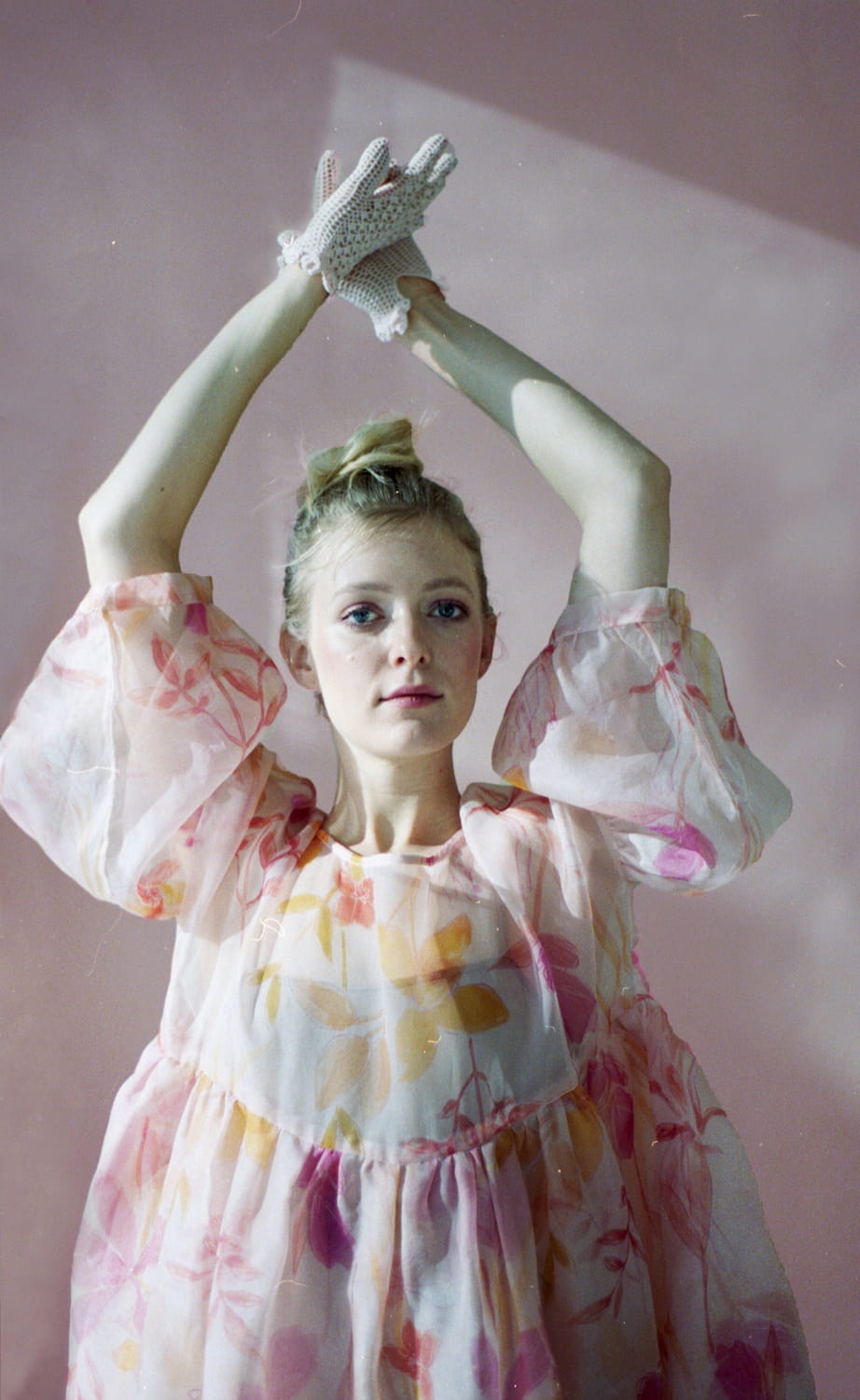 A person is wearing a Céline Floral Dress by Noémiah with sheer puff sleeves crafted from silk organza and lace gloves. Their arms are raised above their head, reminiscent of an Emily Dickinson herbarium, against a soft pink background.