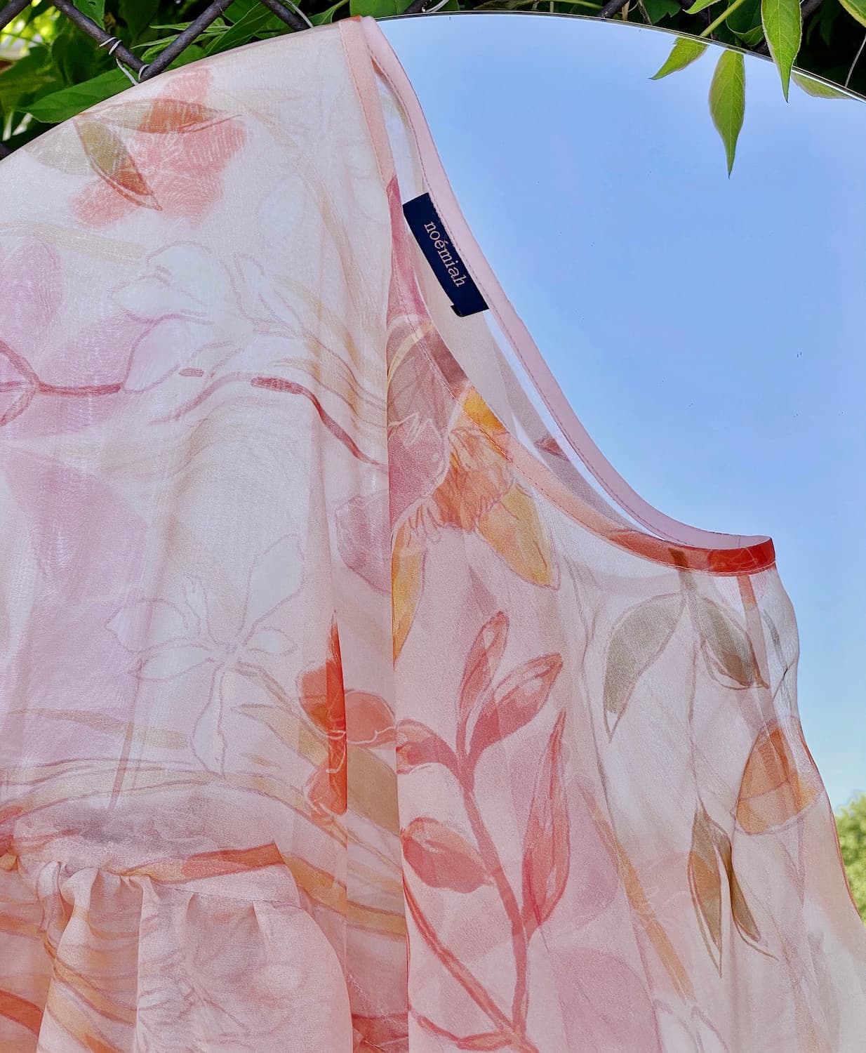 A close-up of a sheer floral dress in soft peach and pink hues, crafted from silk organza, hanging outdoors on a line against a clear blue sky. The neckline reveals a small brand tag reading Noémiah. Green leaves frame the background, reminiscent of an Emily Dickinson herbarium page.
