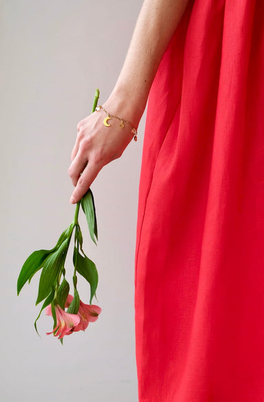 A person in a vibrant red dress holds drooping pink flowers. The arm, adorned with the Jeanne Bracelet by Noémiah, featuring a delicate crescent moon bar chain adorned with Swarovski crystals, subtly glimmers. The background is a simple light gray, emphasizing the dress and the graceful sparkle of gold-plating.