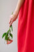 A person in a vibrant red dress holds drooping pink flowers. The arm, adorned with the Jeanne Bracelet by Noémiah, featuring a delicate crescent moon bar chain adorned with Swarovski crystals, subtly glimmers. The background is a simple light gray, emphasizing the dress and the graceful sparkle of gold-plating.