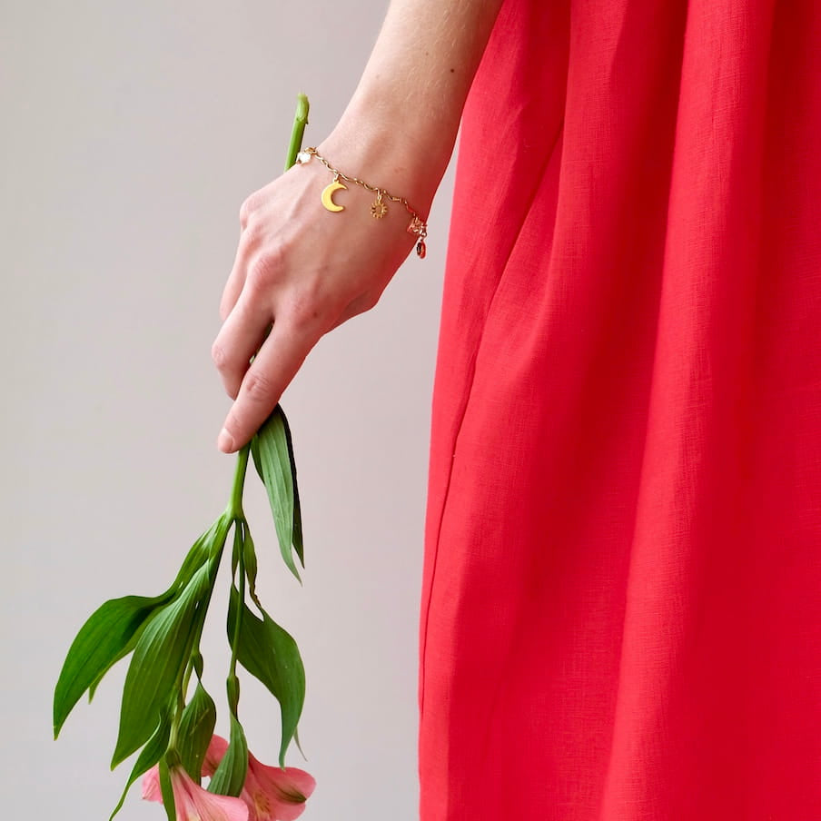 A person in a vibrant red dress holds drooping pink flowers. The arm, adorned with the Jeanne Bracelet by Noémiah, featuring a delicate crescent moon bar chain adorned with Swarovski crystals, subtly glimmers. The background is a simple light gray, emphasizing the dress and the graceful sparkle of gold-plating.