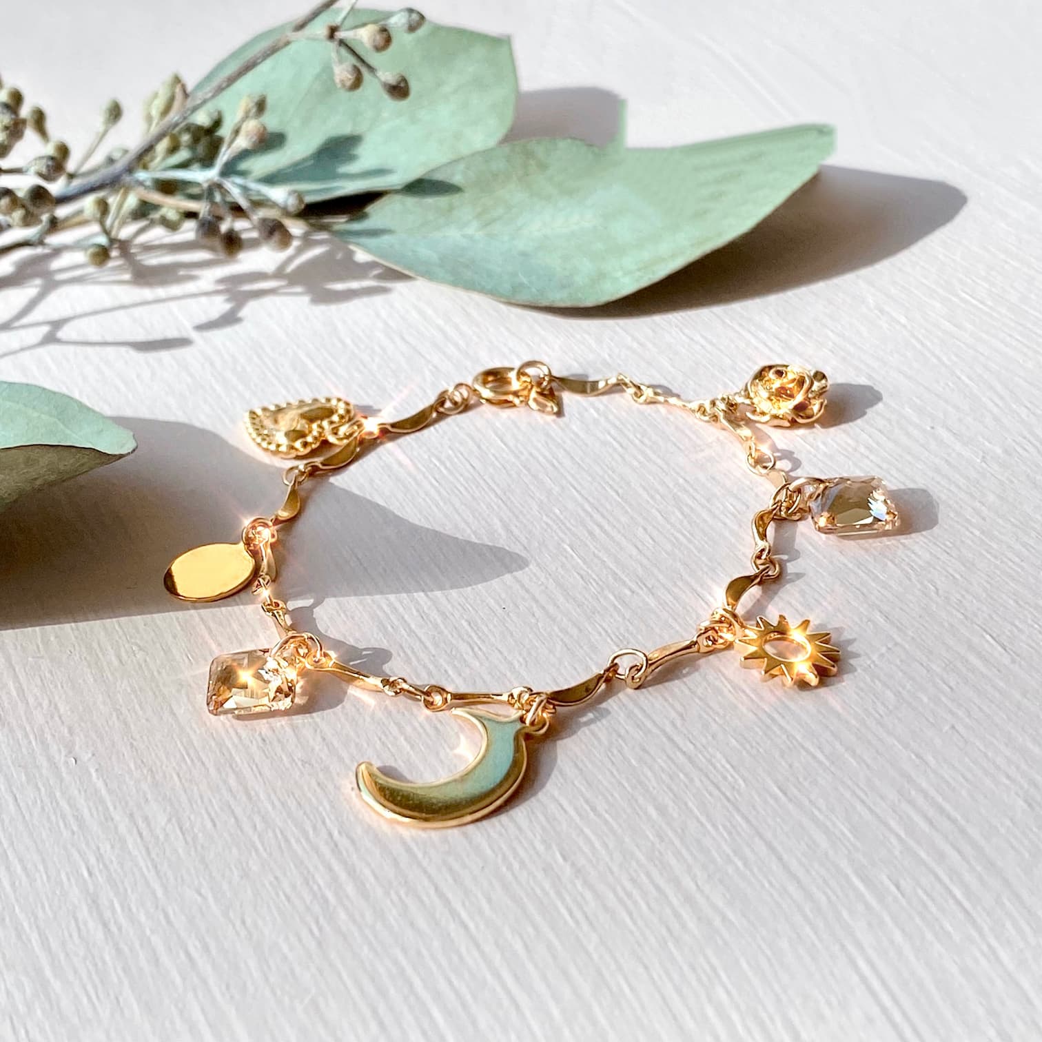 The Jeanne Bracelet by Noémiah, featuring a gold-plated finish with various charms such as a crescent moon, star, heart, and round shapes embellished with Swarovski crystals, is showcased on a light surface. Green leaves are artfully arranged around the bracelet for decoration.