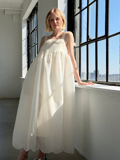 Wearing the Noémiah Jeanne Dress — Bridal Edit, a woman stands by large sunny windows in a bright, minimalist room, softly illuminated by natural light.