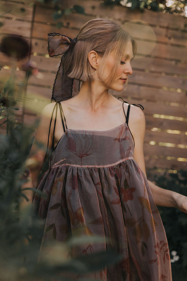 A woman with short blonde hair, wearing a floral dress with scalloped embroidered edges and a large Silk Ribbon from Noémiah’s Floral Edit in her hair, stands outdoors near greenery and a wooden fence, looking down with a calm expression.