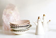 A stack of handmade scalloped-edge ceramic side plates in pastel hues is placed next to two Jewelry Dishes from Noémiah, expertly crafted in the shape of cacti with shimmering gold tips. In the background, a large pink crystal rests on a white surface.