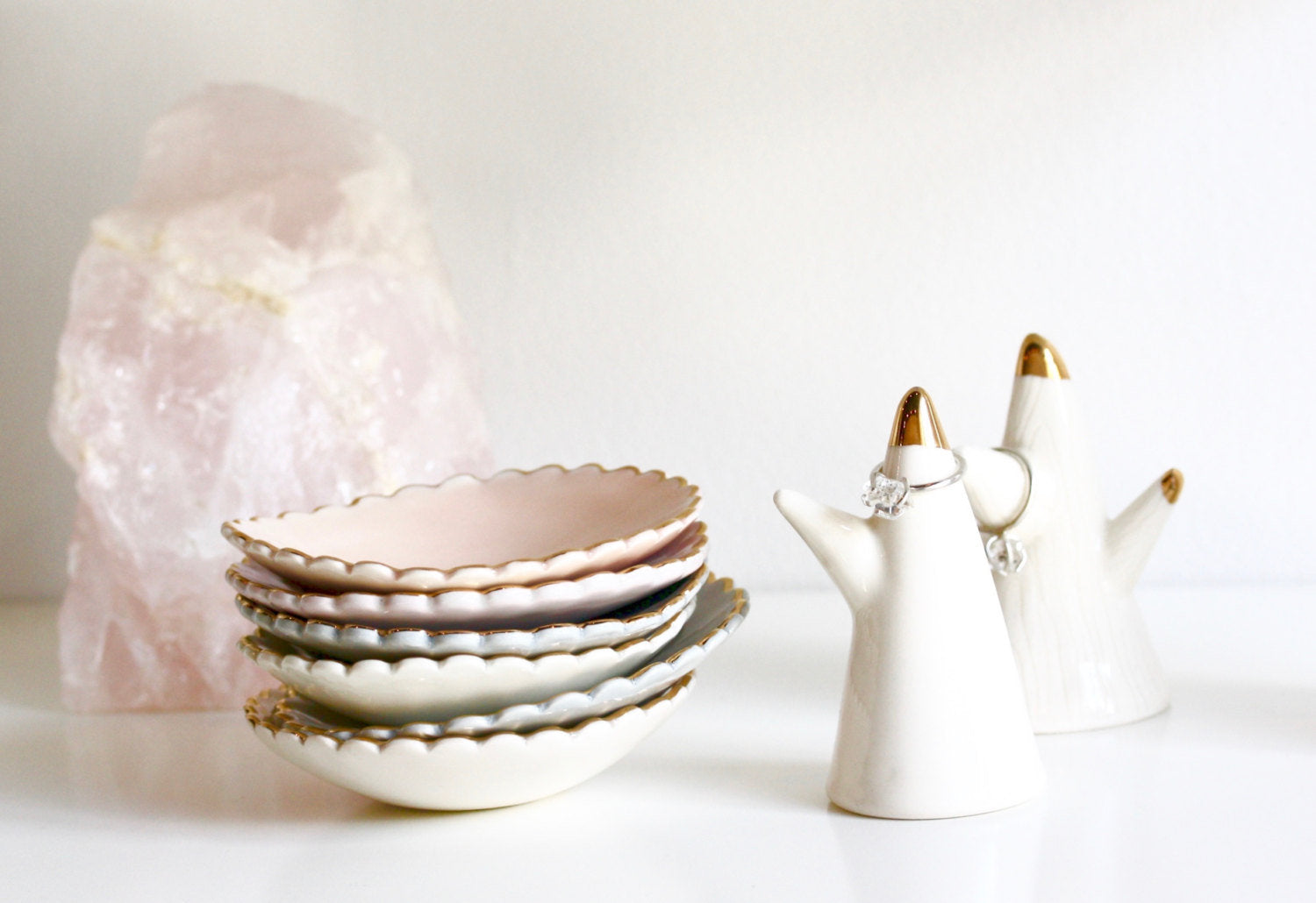 A stack of handmade scalloped-edge ceramic side plates in pastel hues is placed next to two Jewelry Dishes from Noémiah, expertly crafted in the shape of cacti with shimmering gold tips. In the background, a large pink crystal rests on a white surface.