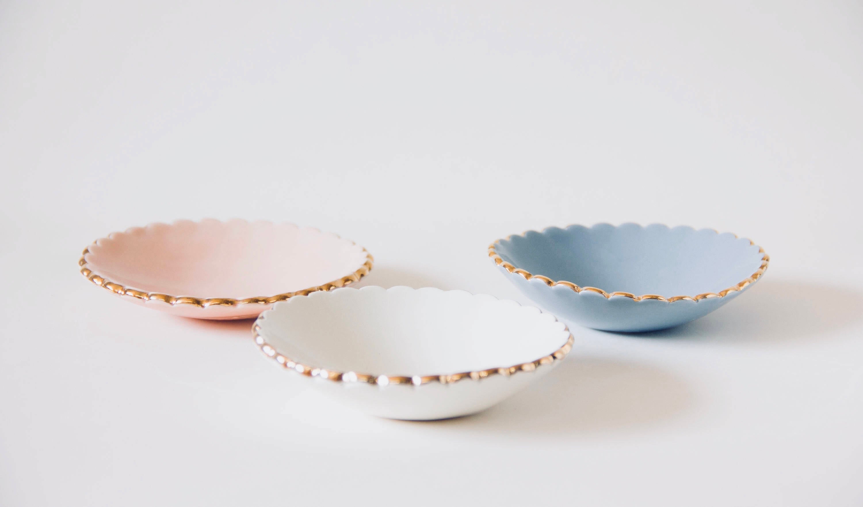 The Noémiah Jewelry Dishes, a set of three small, handmade ceramic bowls featuring scalloped edges in pastel shades of pink, white, and blue, are elegantly arranged on a white surface. Each dish is adorned with a gold-trimmed rim.