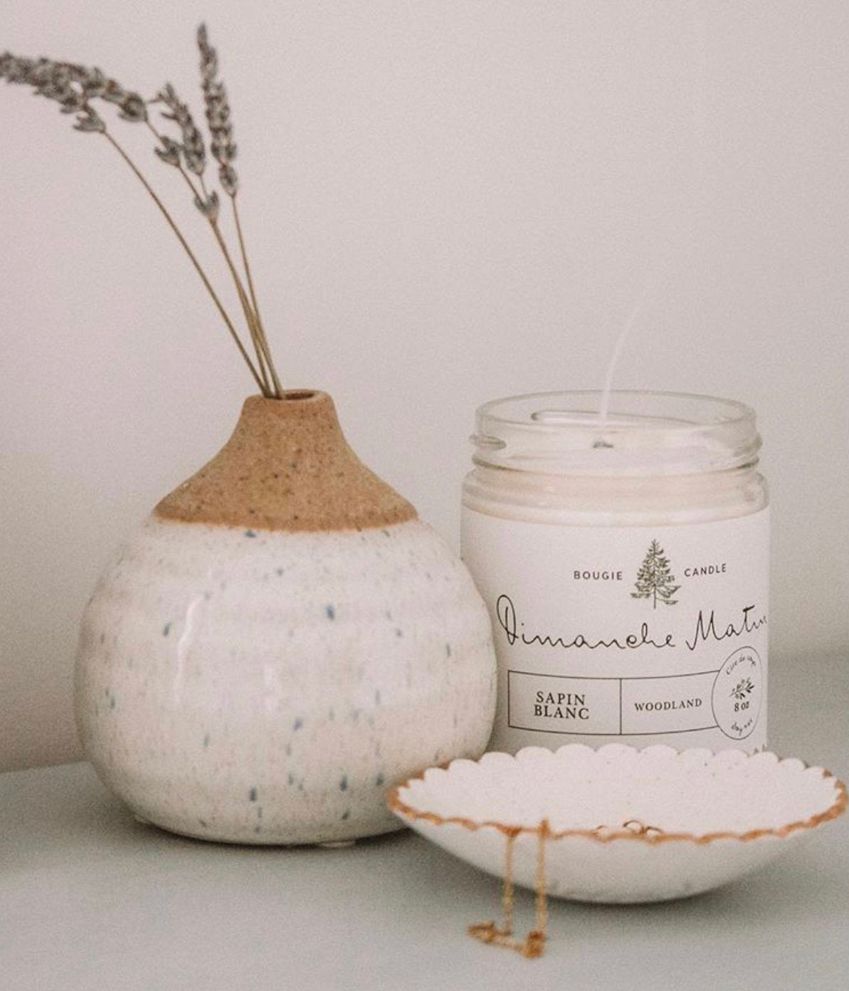 A white speckled ceramic vase filled with lavender stems, a lit candle in a glass jar labeled Dimanche Matin, and Noémiah's handmade Jewelry Dish featuring gold pieces adorn a light surface.