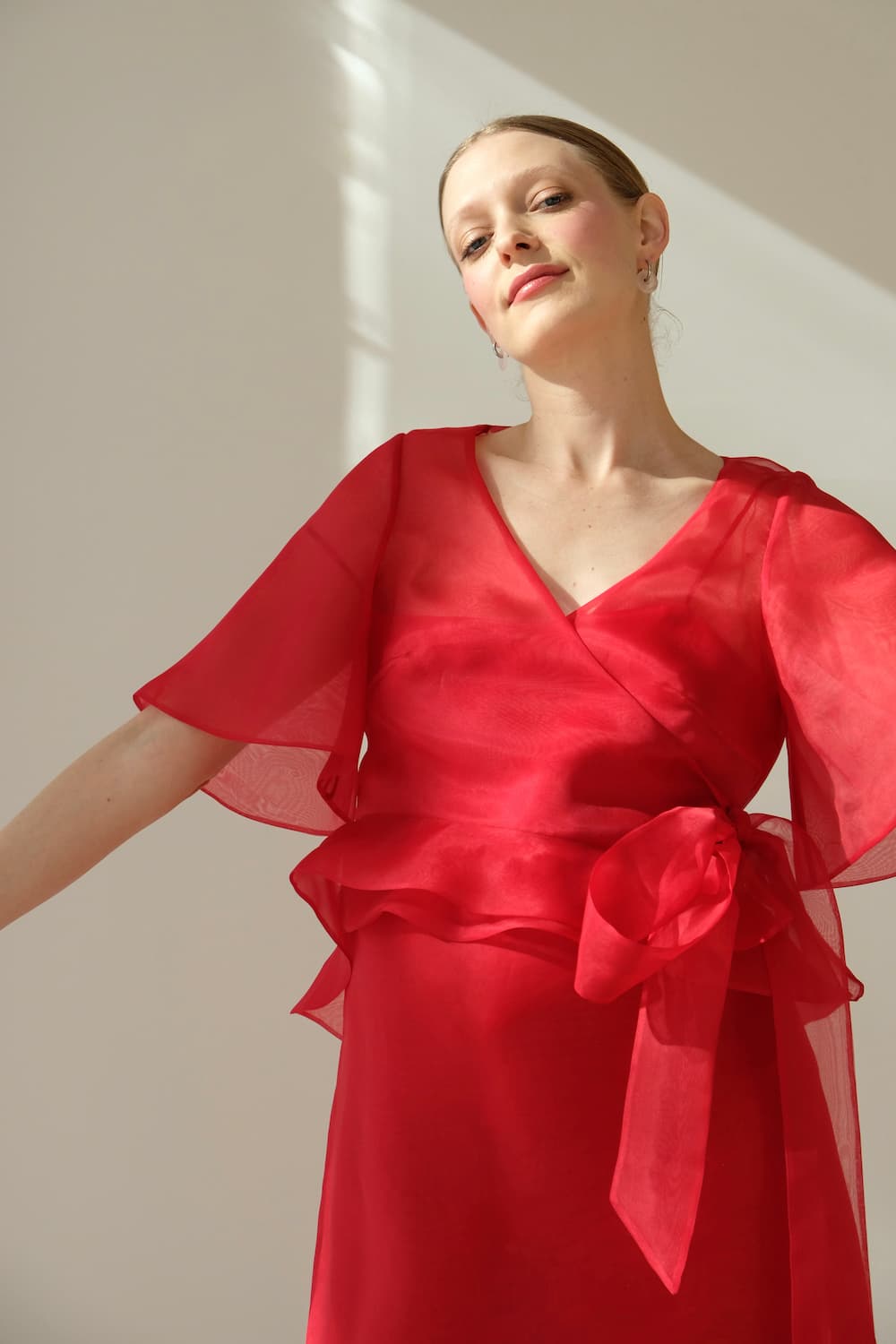 A woman confidently poses against a light background, wearing the red, sheer Julia Wrap Top by Noémiah, complete with flutter sleeves and a large ribbon.