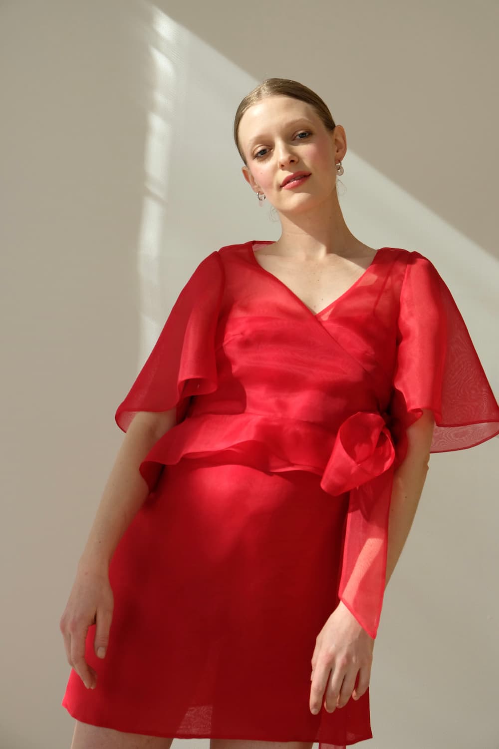 A person stands against a light background wearing the Noémiah Julia Wrap Top, featuring scalloped puffed sleeves and decorative waist detail. They have light hair pulled back, looking toward the camera as soft shadows are cast by the lighting.