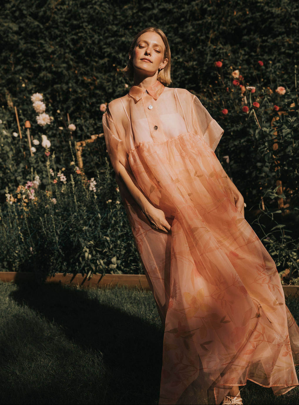A woman stands in a sunlit garden surrounded by greenery and blooming flowers, wearing the June Shirt Dress by Noémiah—a flowing, sheer peach-colored silk organza dress with short sleeves and buttons.