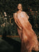 A woman stands in a sunlit garden surrounded by greenery and blooming flowers, wearing the June Shirt Dress by Noémiah—a flowing, sheer peach-colored silk organza dress with short sleeves and buttons.