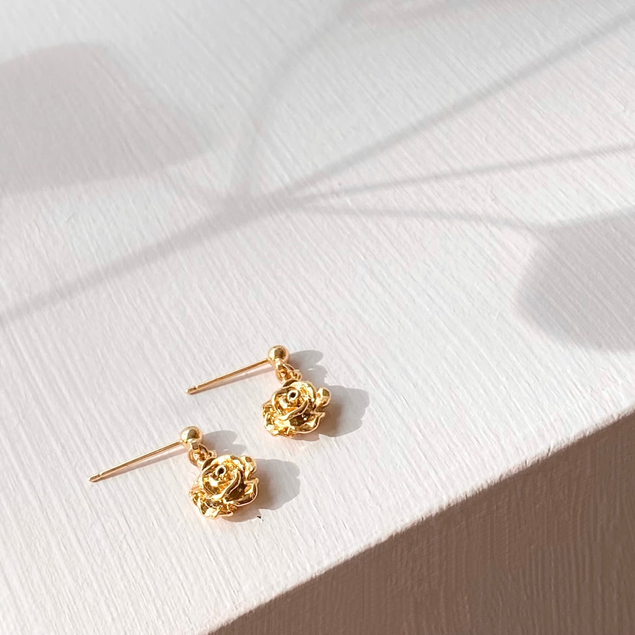 A pair of Justine Earrings by Noémiah, featuring gold-plated rose-shaped studs, is displayed on a textured white surface. The light casts soft shadows that enhance the warm and elegant atmosphere, making these stunning accessories ideal for those with sensitive ears.