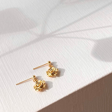 A pair of Justine Earrings by Noémiah, featuring gold-plated rose-shaped studs, is displayed on a textured white surface. The light casts soft shadows that enhance the warm and elegant atmosphere, making these stunning accessories ideal for those with sensitive ears.