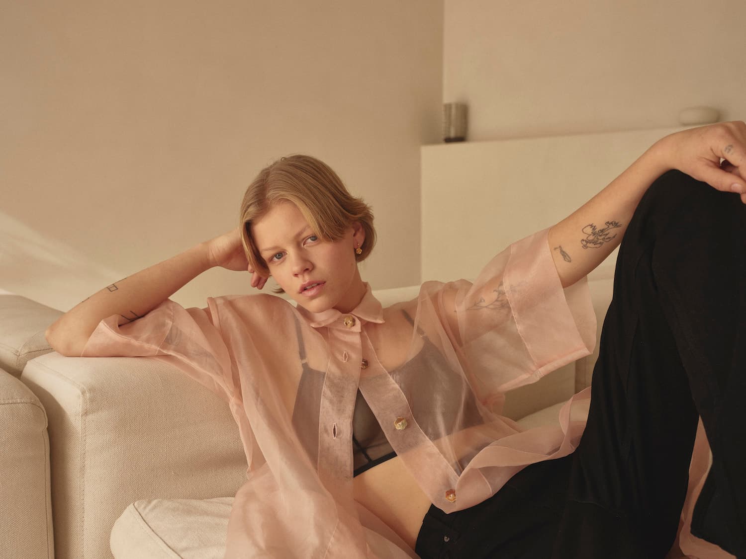 A person with short hair lounges on a white sofa, wearing a sheer pink button-up shirt over a black top and black pants. The Noémiah Justine Earrings catch the soft light in the room with neutral-toned walls, adding a subtle touch of elegance to the relaxed and minimalist atmosphere.