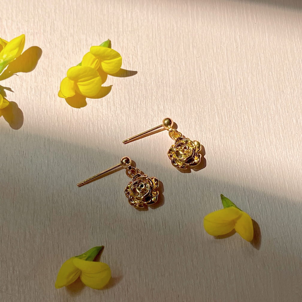 The Justine Earrings by Noémiah, crafted in a gold-plated flower shape, rest on a light wooden surface surrounded by small yellow flowers. The sunlight casts soft shadows, creating a warm and inviting atmosphere. These clip-on earrings are perfect for sensitive ears, offering both style and comfort without compromising elegance.