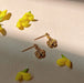 The Justine Earrings by Noémiah, crafted in a gold-plated flower shape, rest on a light wooden surface surrounded by small yellow flowers. The sunlight casts soft shadows, creating a warm and inviting atmosphere. These clip-on earrings are perfect for sensitive ears, offering both style and comfort without compromising elegance.