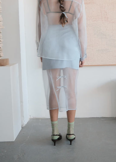 In a minimal, modern setting, a person with braided hair wears the Noémiah Leah Skirt over a light blue slip and sheer silk organza long-sleeve dress, styled with green socks and black heels, facing away indoors.