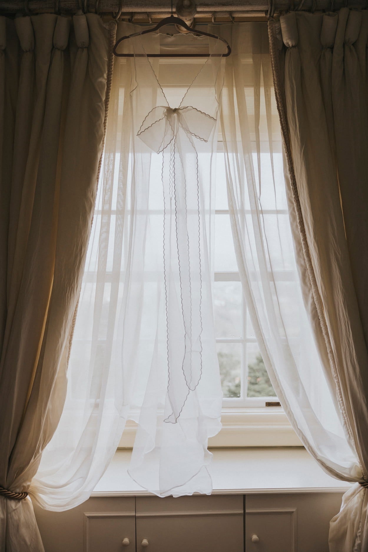 The Lia Dress by Noémiah, crafted from sheer white silk organza and adorned with delicate embroidery, gracefully drapes across the window, softly diffusing the natural light. Tied back on both sides, it creates a symmetrical and elegant look reminiscent of a minimal wedding dress for non-traditional brides.