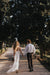 A couple strolls hand in hand down a sunlit path edged with trees. The woman, embracing her unique style as a bride, is dressed in Noémiah's Lia Dress made of silk organza and holding a bouquet, while the man wears a white shirt paired with black pants. Shadows and greenery envelop them in this serene outdoor setting.