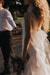 A couple holds hands during a romantic outdoor moment. The woman, wearing the Lia Dress by Noémiah, carries a bouquet of flowers. Sunlight casts soft shadows, highlighting her curly hair. The man's face is not visible, adding a sense of mystery to this enchanting scene featuring non-traditional brides.