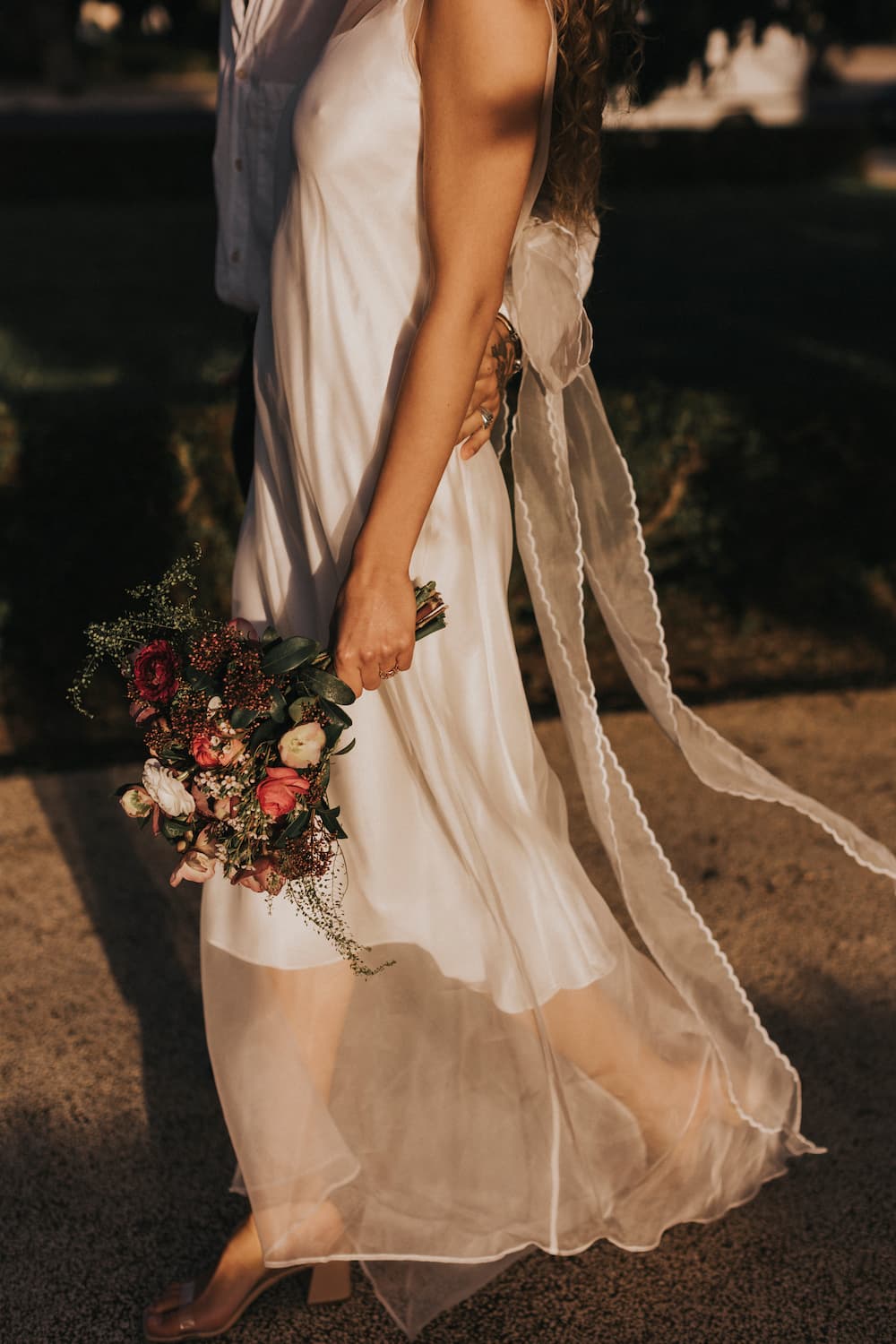 A woman in the elegant Lia Dress from Noémiah, crafted from silk organza with a sheer overlay, holds a bouquet of pink and white flowers while walking along a sunlit path. She wears clear-heeled shoes, creating a romantic and warm ambiance ideal for non-traditional brides.