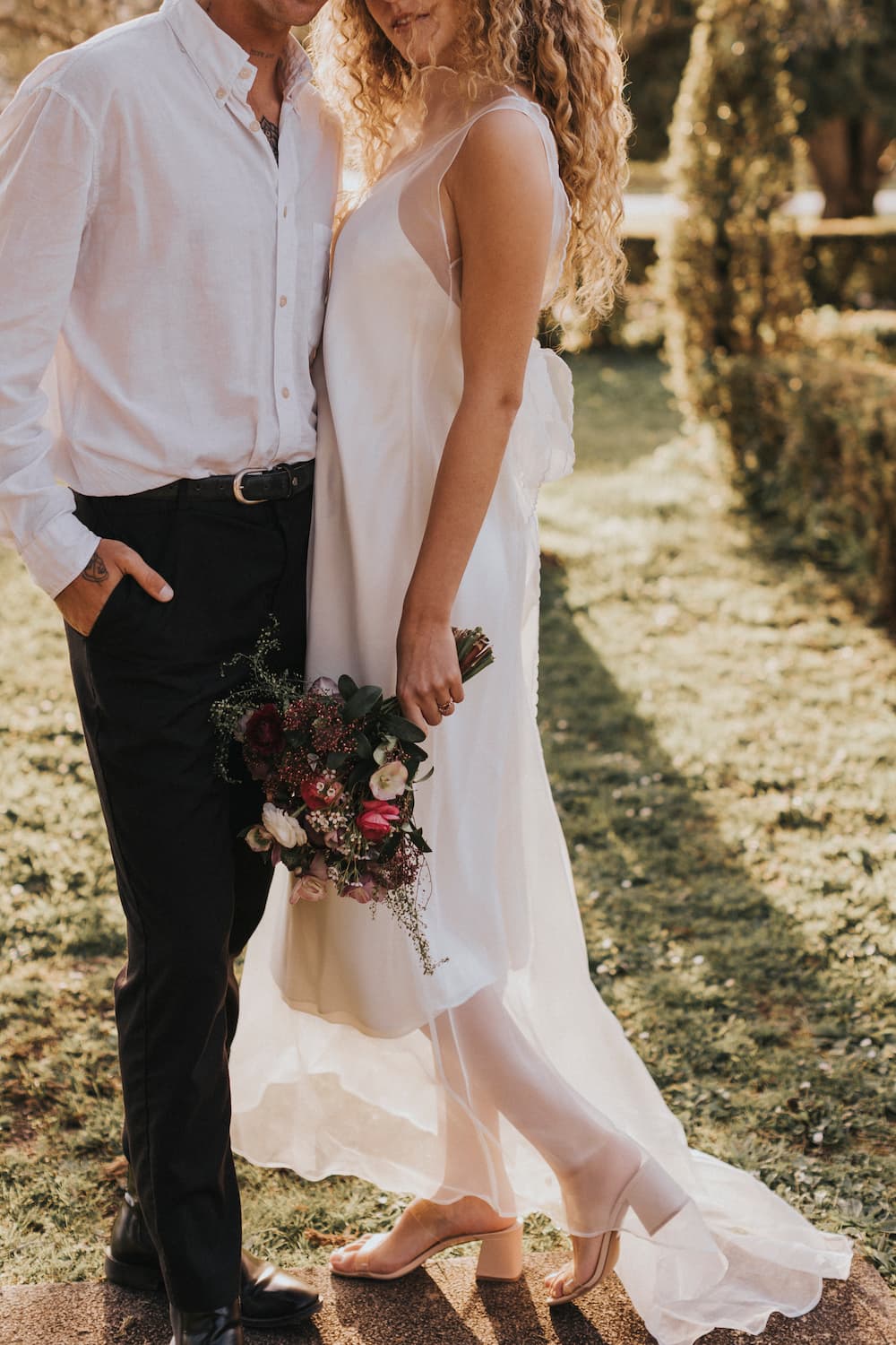 A couple stands closely in a garden, radiating elegance in their formal attire. The woman, a non-traditional bride, wears the minimal and chic Lia Dress by Noémiah while holding a bouquet of flowers. The man complements her look in a white shirt and dark pants with his hand in his pocket, savoring the sunlight casting shadows on the grass.